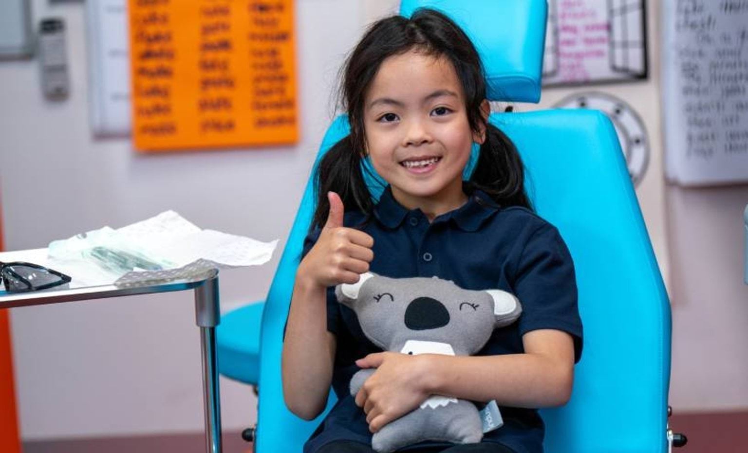 Girl sitting in a turquoise dentist chair giving a thumbs up and hugging a koala bear toy