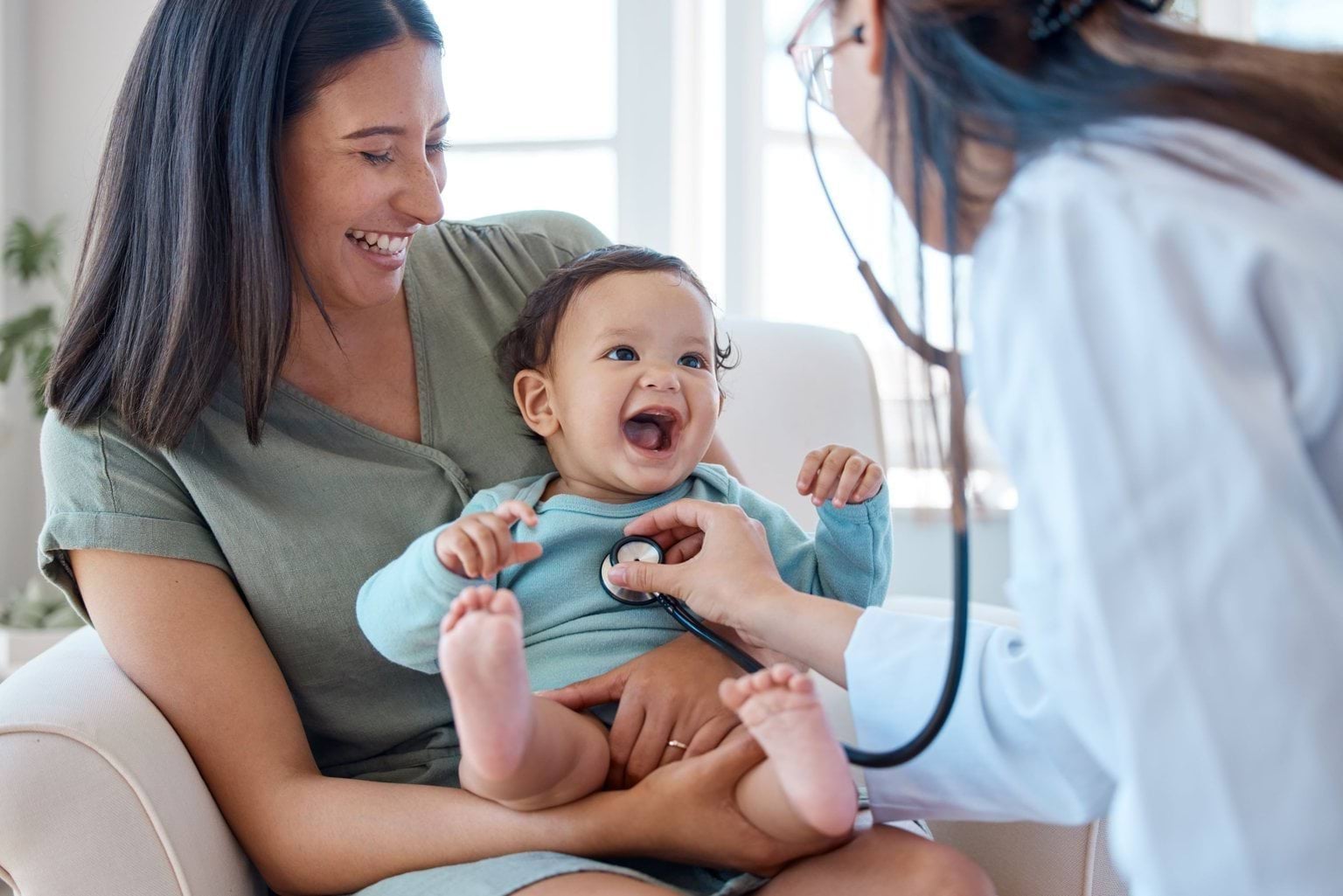 A baby laughs in the lap of his Mum while GP checks his heart