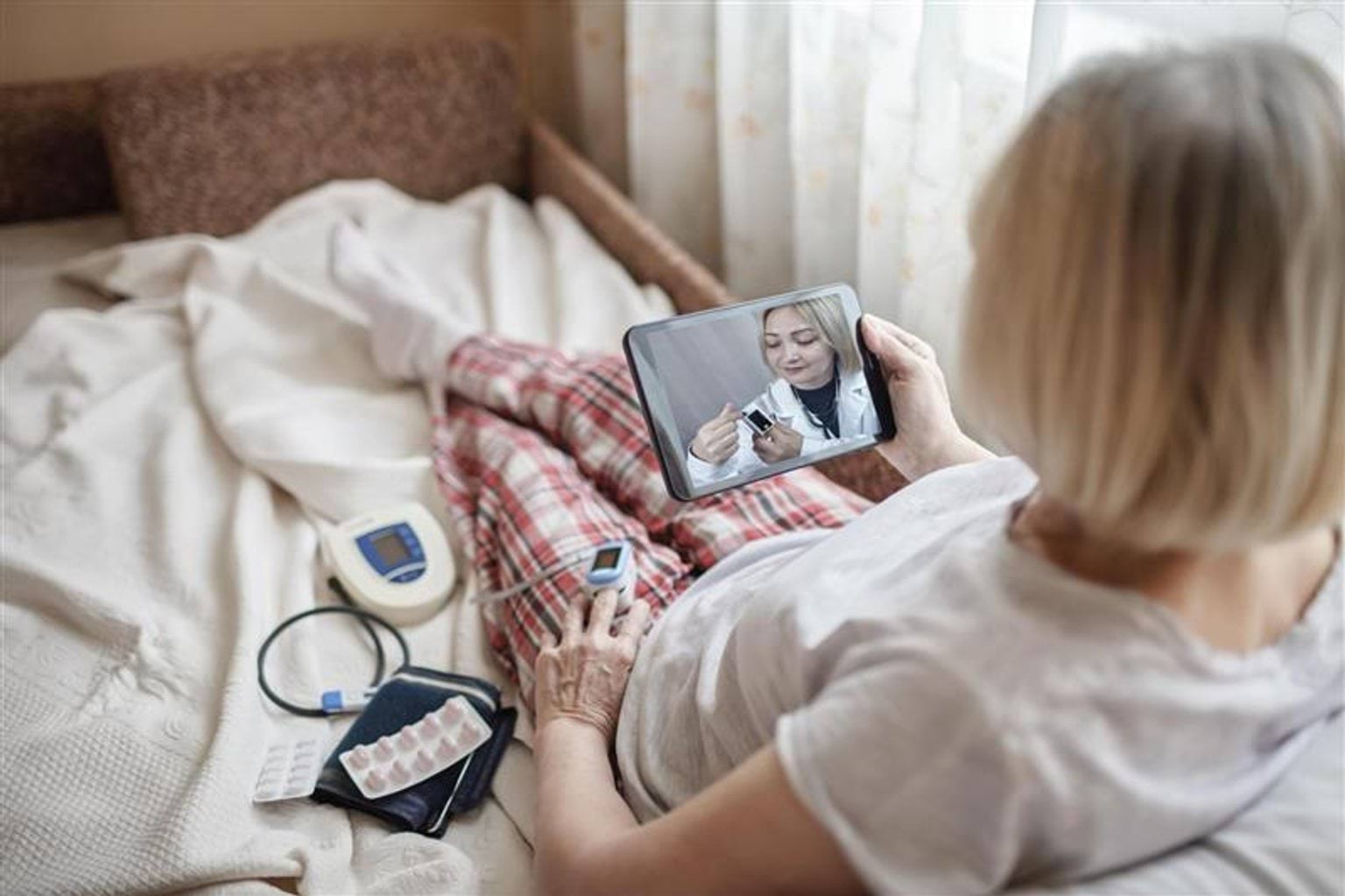 Person lying on a bed wearing plaid pyjama pants, holding a tablet showing a healthcare professional during a telehealth consultation as part of the Virtual Hospital pilot. A blood pressure monitor, a pulse oximeter clipped to one finger, and several blister packs of medication are placed nearby.