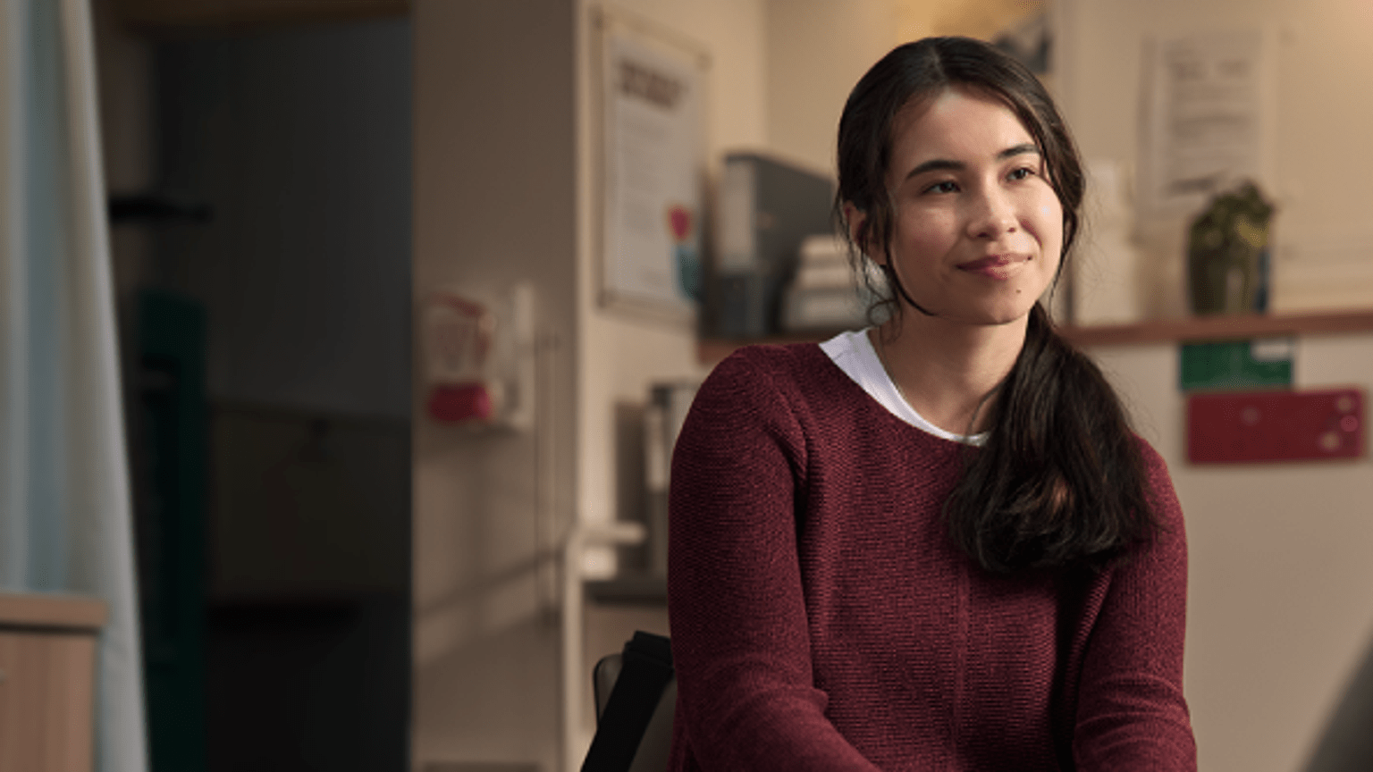 Person with hair tied back, wearing a maroon sweater, seated in a clinical consultation room with medical equipment and posters visible in the background. The person is turned slightly to the side, with hands together, in soft indoor lighting.
