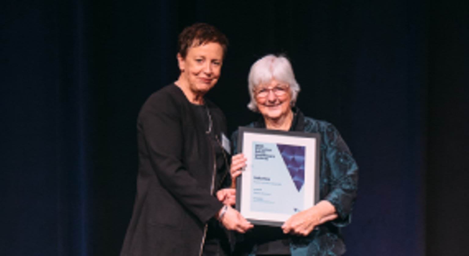 Photograph of two people standing on a stage and holding an award certificate at the Victorian Public Healthcare Awards.