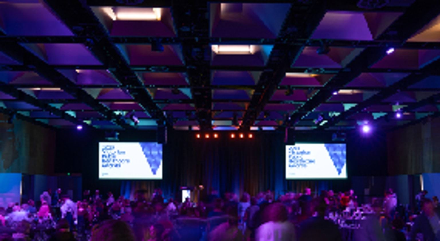 Photograph of a the Victorian Public Healthcare Awards. Attendees are seated at round tables facing a stage with a lectern and large presentation screens on either side.