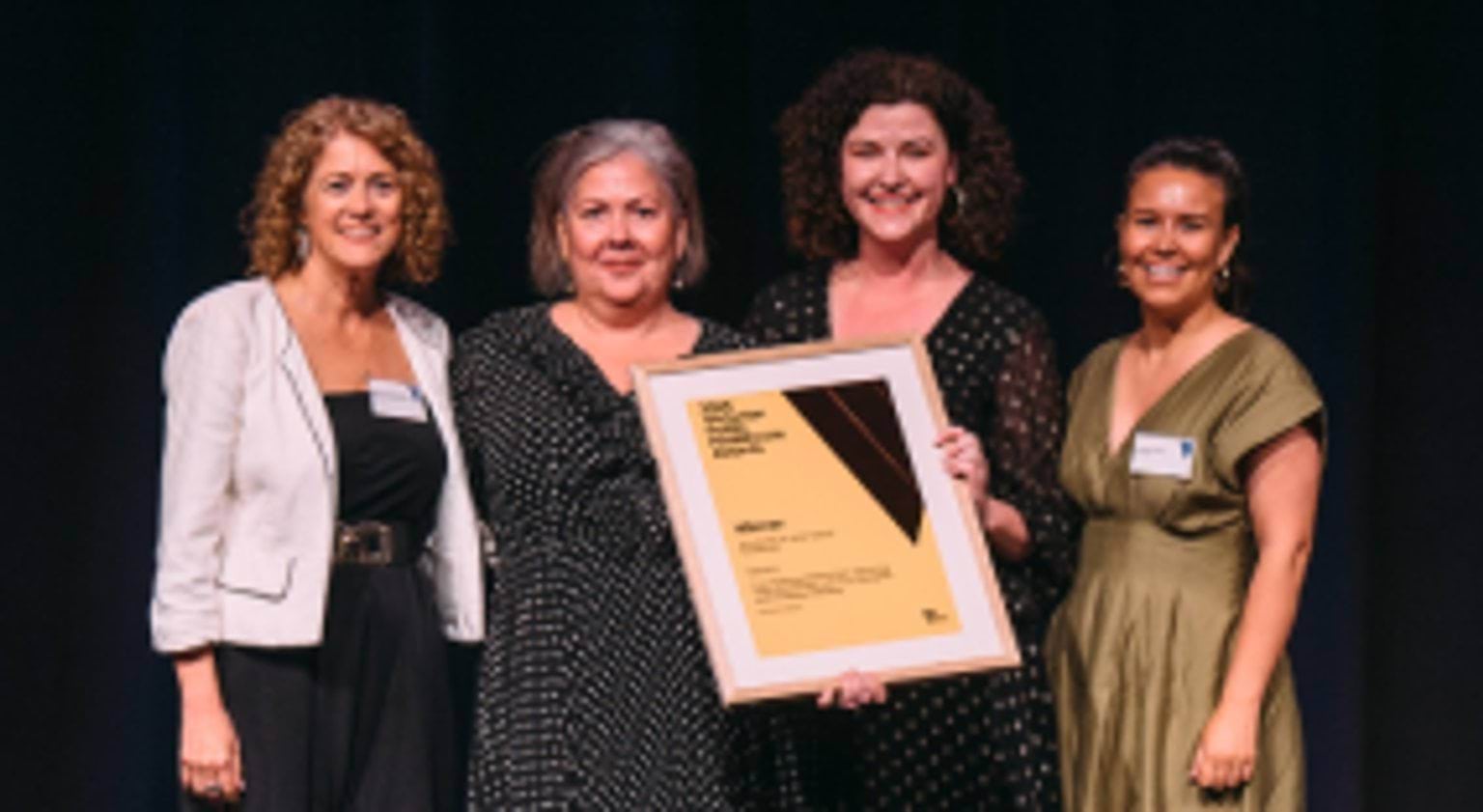 Photograph of four people standing on a stage and posing with a framed award certificate at the 2025 Victorian Public Healthcare Awards