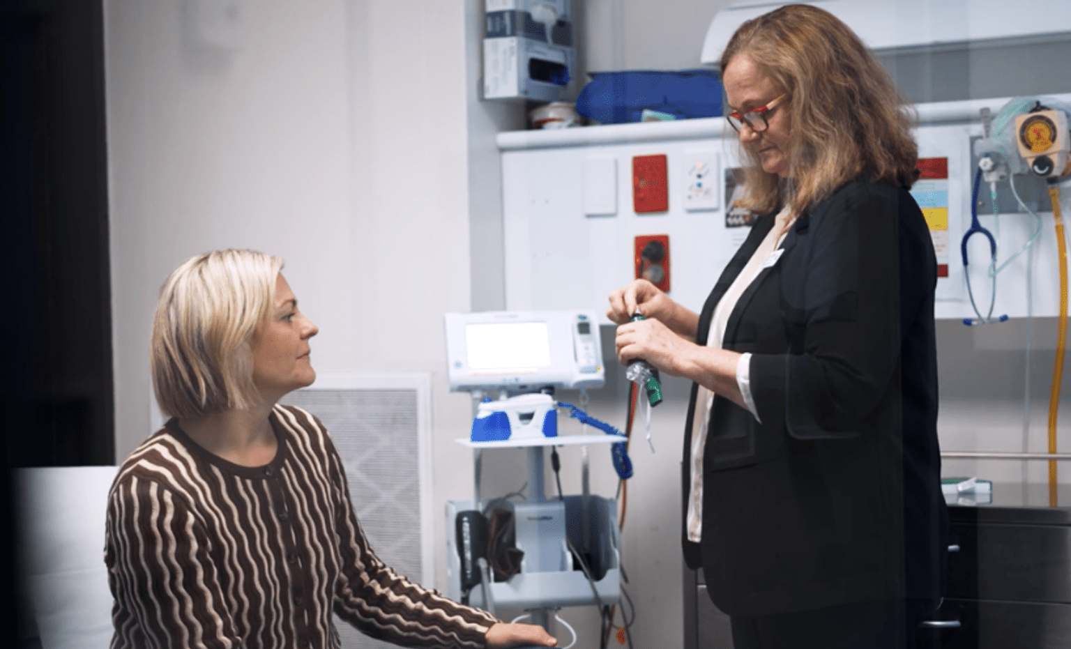 Healthcare consultation in a clinical setting with two women and medical equipment.