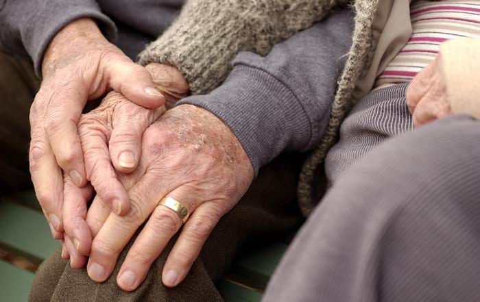 The photo is of hands of two older people, male and female, resting on the knee of the man. It demonstrates intimacy and comfort between the two people.
